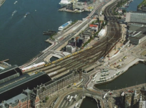 Luchtfoto van de De Ruijterkade in Amsterdam met het Centraal Station, treinsporen, tram- en busstations en het IJ op de achtergrond.